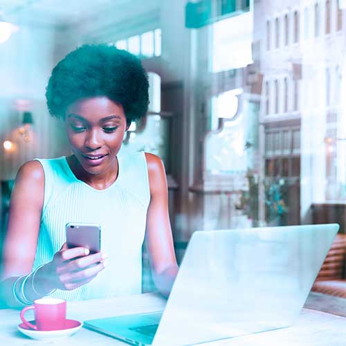 Woman using a smartphone while working on a laptop at a table with a cup of coffee.