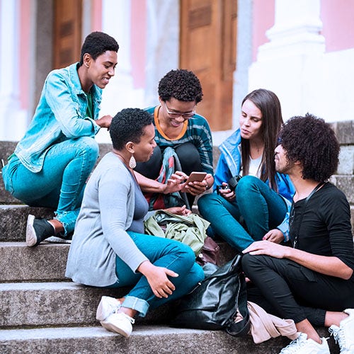 Group of five young adults sitting on steps together, looking at a phone and chatting.