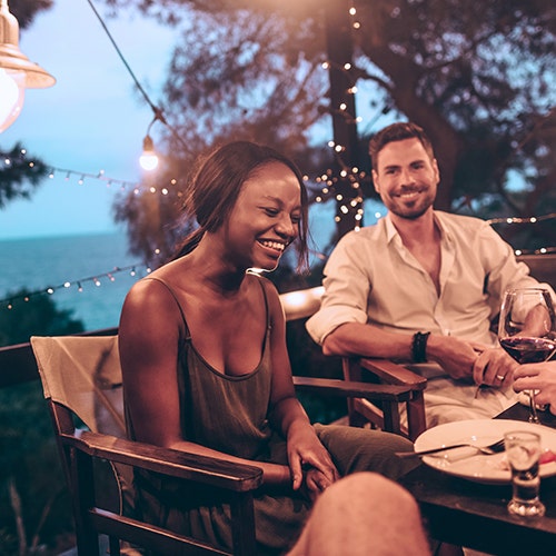 Two people sitting at an outdoor table at dusk, smiling and enjoying drinks with string lights in the background.