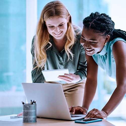 Two women smiling while reviewing information on a smartphone and laptop together in a bright office.