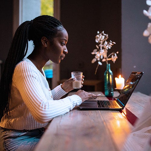 Woman working on a laptop at a table with a candle and drink, focused in a dimly lit setting.