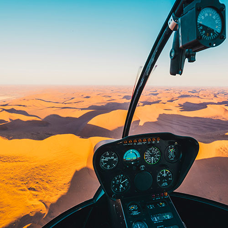 View from inside a helicopter cockpit flying over expansive desert dunes under a clear sky.