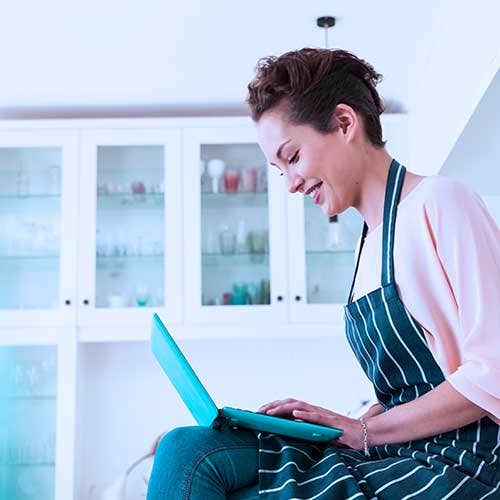 Woman sitting in a bright kitchen wearing an apron, smiling while using a laptop.